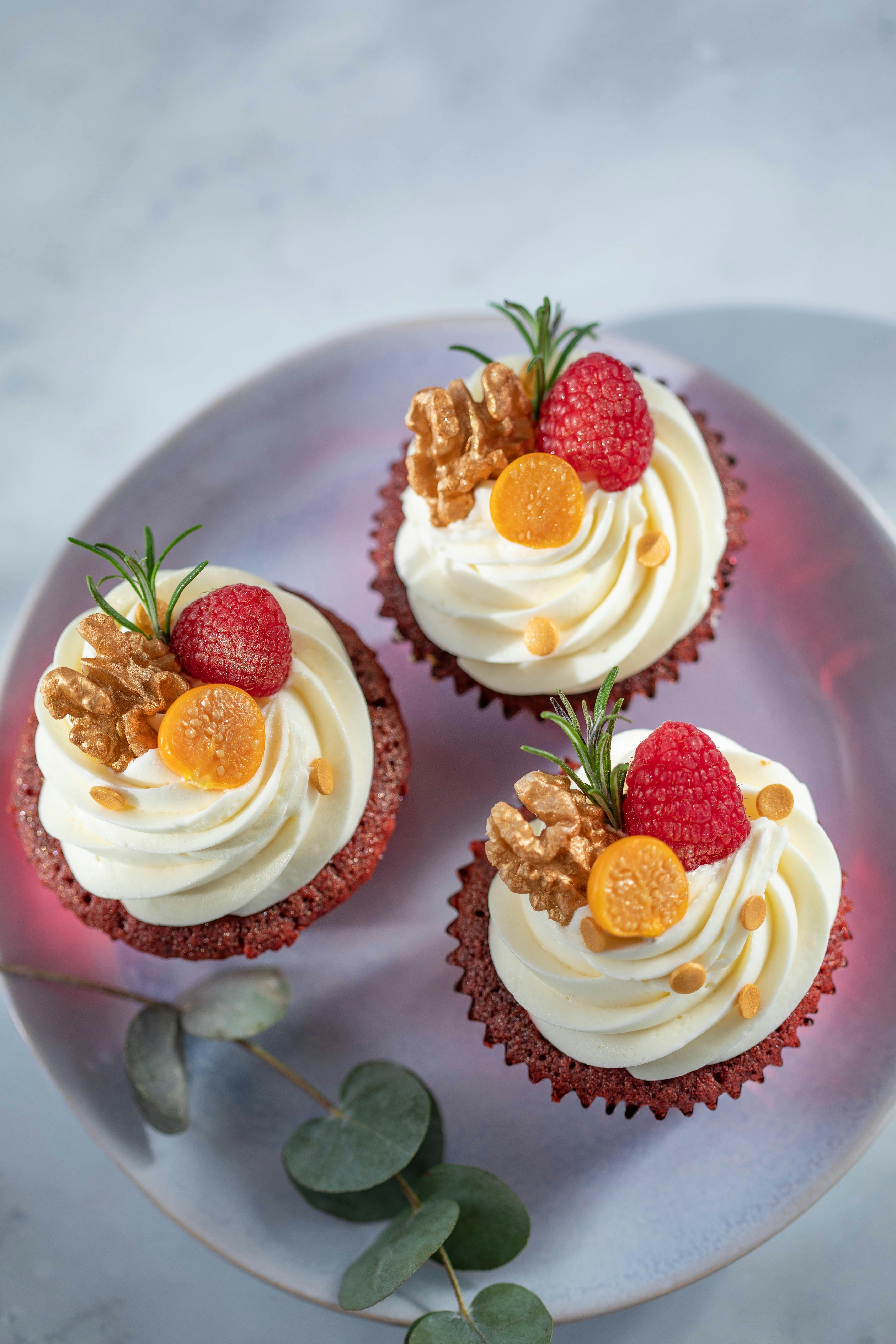 Three red velvet cupcakes with cream cheese frosting, raspberries and walnuts